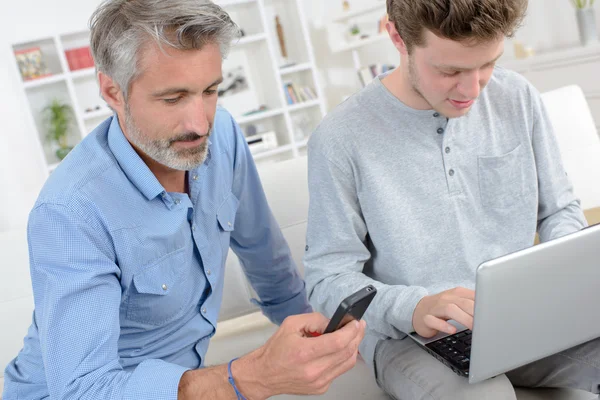 Giovane sul computer portatile, uomo più anziano utilizzando smartphone — Foto Stock