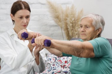 Terapist, yaşlı kadına egzersizlerde yardımcı oluyor. Fizik tedavi seansında halterleri kullanan yaşlı bir hasta. Rehabilitasyon sağlık hizmeti kavramı.