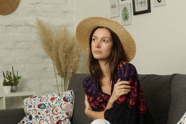 Boho tarzında sıradan bir portre. Hasır şapka takan genç bir kadın, evinde kanepede oturuyor..