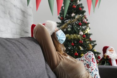 Noel Baba şapkası takan ve koruyucu yüz maskesi takan bir kadının Noel ağacının yanındaki koltukta dinlendiği yakın plan. Corona virüs salgını sırasında kış tatili, kendini karantina konsepti. 