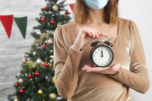 サンタ帽の女性と大晦日に目覚まし時計を保持保護顔マスク コロナウイルス発生中の冬の休日 自己検疫の概念 — ストック写真