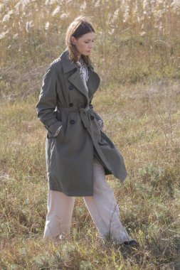 Açık havada beyaz kadın portresi. Geniş bacaklı, bej pantolonlu ve yeşil trençkotlu..