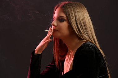Esmer ve esmer bir kadının stüdyo portresi. Arka planda sigarası var. Kameraya bakıyor.
