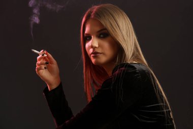 Esmer ve esmer bir kadının stüdyo portresi. Arka planda sigarası var. Kameraya bakıyor.