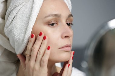 Genç bir kadın aynada yüzünü ve deri dokusunu inceliyor.