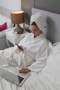 Genç beyaz kadın yatakta cep telefonu ve dizüstü bilgisayar kullanıyor. 