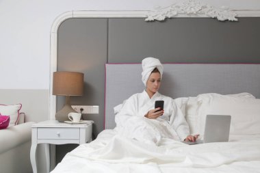 Genç beyaz kadın yatakta cep telefonu ve dizüstü bilgisayar kullanıyor. 