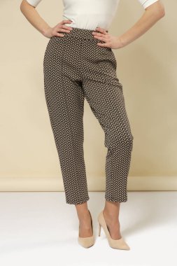 Stüdyo fotoğrafçılığı. Rahat sigara içen, kahverengi desenli kırpık pantolon giyen ve yüksek topuklu ayakkabı giyen genç bir kadın model.