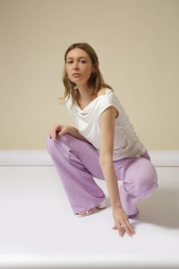Kızaksız beyaz pamuklu gömlek ve leylak keten pantolon giymiş genç bir modelin stüdyo fotoğrafları. Gündelik ve şık kıyafetler, bahar-yaz moda trendi.