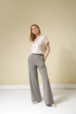 Açık gri, şık, gündelik, yüksek kaliteli pantolon ve V yaka beyaz bluz giyen kadın model. Zarif, basit bir iş görünüşü. Stüdyo çekimi.