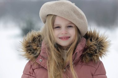 Açık havada 8 yaşındaki sevimli kızın portresi bej renkli yün bereli ve tüylü pembe kışlık ceketli.