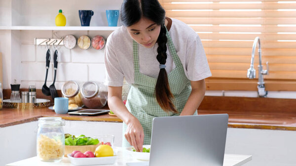 Cooking at home, Asian woman looking on the digital recipe while cooking healthy meal, Asia girl preparing food by following food blog on laptop computer in the kitchen, E learning homemade food concept 