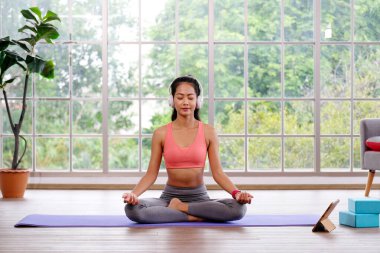 Evde yoga meditasyonu yaparken kulaklık takan Asyalı kadın, sabahları rahat yoga pozisyonu için mindere oturan genç Asyalı kadın, evde egzersiz, refah, akıl sağlığı...