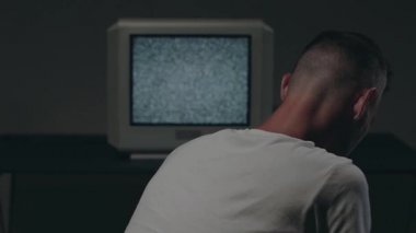 Beyaz tişörtlü adam Eski Gümüş TV karşısında oturuyor.