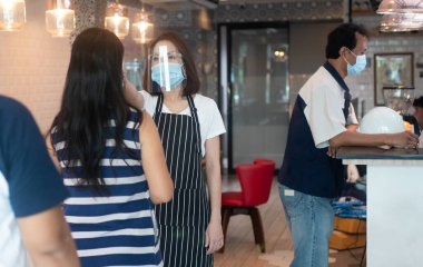 Asyalı garson kadın yüz maskesi takıyor ve elinde kızılötesi alın termometresi var. Restorana girmeden önce vücut ısısını ölçmek için müşterilerin semptomlarını kontrol ediyor. ).