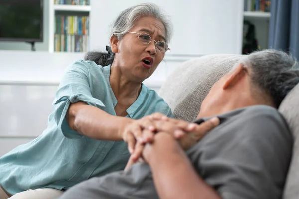 Asian old elderly man is In pain with his hands on his chest and having a heart attack in the Living room and his wife is shocked and confused, helpless. Concept of healthcare and Elderly caregivers