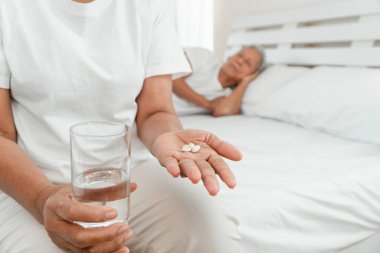 Yatak odasındaki yatağın üzerinde elinde ilaç ve su tutan yaşlı bir kadın ve kocası yanında uyuyor. Yaşlılarda uyku sorunları, yatıştırıcı kullanımı ve uyku ilacı bağımlılığı kavramı