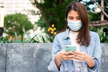 Anti-koronavirüs COVID-19 salgın hastalıkları için maske takmış genç bir kadının portresi ve halka açık alanda akıllı telefon kullanımı. Virüs salgını ve kirliliği kavramı