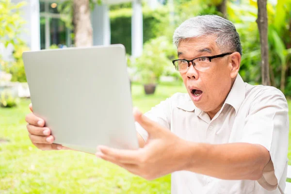 Portrait of an old elderly Asian man is holding a computer laptop and surprised after got some email and read some news in the backyard. Concept of no Ageism and Hobbies after retirement.