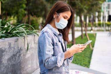 Anti-koronavirüs COVID-19 salgın hastalıkları için maske takmış genç bir kadının portresi ve halka açık alanda akıllı telefon kullanımı. Virüs salgını ve kirliliği kavramı
