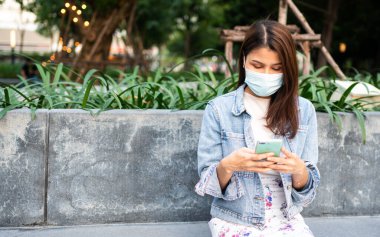 Anti-koronavirüs COVID-19 salgın hastalıkları için maske takmış genç bir kadının portresi ve halka açık alanda akıllı telefon kullanımı. Virüs salgını ve kirliliği kavramı