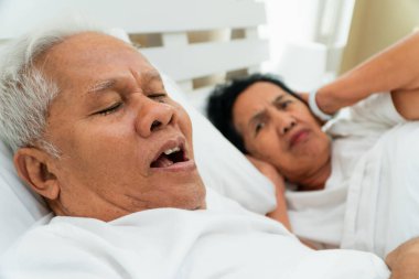 Older Asian woman laying open her eyes and Cover ears with her hands in bed beside her husband who snores and Makes noise, Marital problems. clipart
