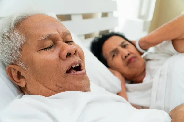 Older Asian woman laying open her eyes and Cover ears with her hands in bed beside her husband who snores and Makes noise, Marital problems.