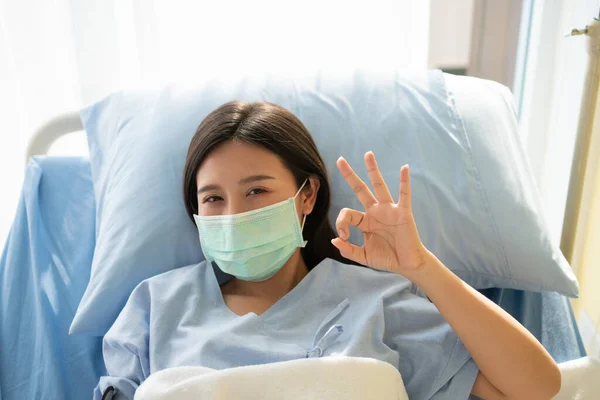 Happy Asian Female Patient Wears Mask Lies Bed Shows Symbol — Foto Stock