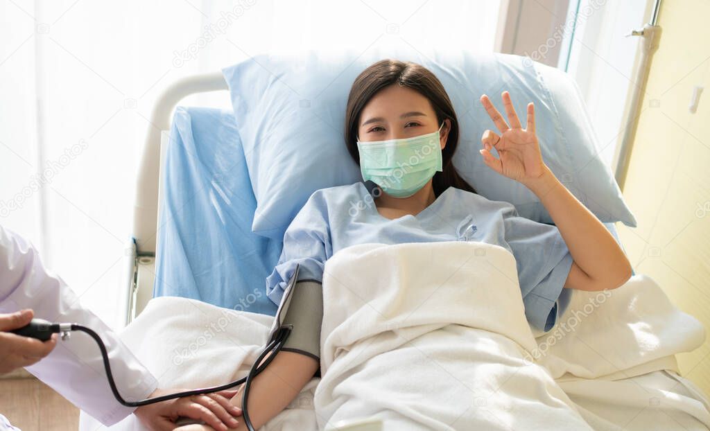 Happy Asian female patient wears a mask, lies on the bed, and Shows an ok symbol. When the doctor examines the symptoms using a pressure gauge. Concept of believe in treatment And insurance coverage