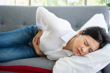 Mutsuz Asyalı kadın kanepede oturuyor ve karın ağrısı çekiyor. Adet, ishal ya da hazımsızlıktan kaynaklanan karın ağrısı. Hastalık ve sağlık hizmeti kavramı