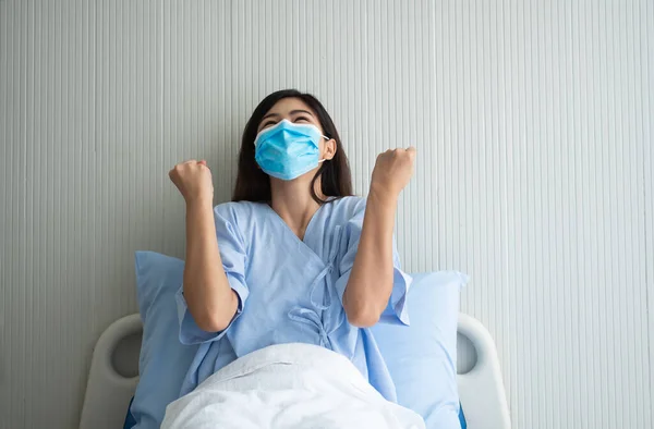 Happy Asian female patient wearing a mask, lies on the bed, and I raised an arms and was delighted for show confidence in treatment. Concept of believe in treatment And insurance coverage