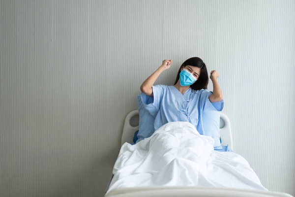 Happy Asian female patient wearing a mask, lies on the bed, and I raised an arms and was delighted for show confidence in treatment. Concept of believe in treatment And insurance coverage