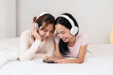 İki birey bir yatağa uzanıyor, gülümsüyor ve kulaklık takıyor, müzik paylaşıyorlar. Samimi ortam ve neşeli etkileşim, evdeki arkadaşlık, bağ ve dijital yaşam tarzını vurguluyor.