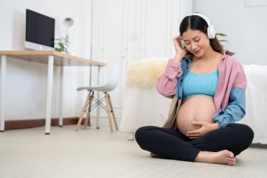Hamile kadın rahat, modern bir evde oturmuş, kulaklıkla müzik dinliyor, karnına hafifçe dokunuyor, annelik bağı kuruyor, duygusal sağlığını koruyor, doğum öncesi bakım, yaşam tarzı, annelik, rahatlama.