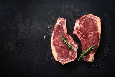 İki çiğ kurutulmuş biftek (cote de boeuf) ve koyu arkaplanda biberiye, biber ve tuz.