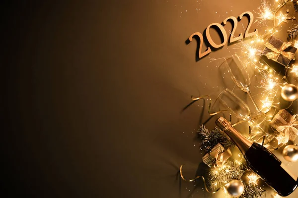 New Years Eve holiday background with fir branches, number 2022, champagne bottle, christmas balls, gift box and lights on dark board