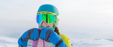 Bir snowboardcu dağların panoramasında bir yamaçta poz veriyor. Dağdan inmeye hazırlanıyor..
