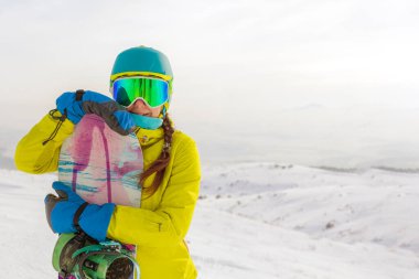 Bir snowboardcu dağların panoramasında bir yamaçta poz veriyor. Dağdan inmeye hazırlanıyor..
