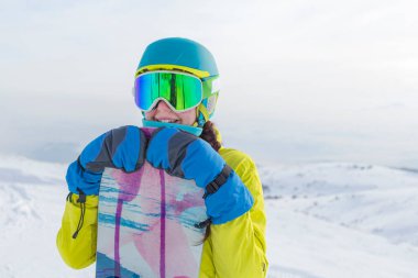 Bir snowboardcu dağların panoramasında bir yamaçta poz veriyor. Dağdan inmeye hazırlanıyor..
