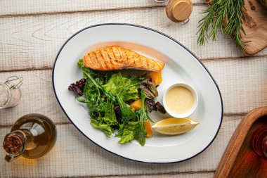 Sulu ızgara somon fileto taze yapraklı yeşillikler ve beyaz tabakta kremalı sosla servis edilir.