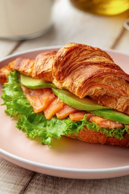 Taze kruvasan sandviçi füme somon, avokado dilimleri ve pembe tabakta servis edilen marul ile dolu.