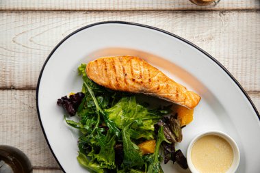 Sulu ızgara somon fileto taze yapraklı yeşillikler ve beyaz tabakta kremalı sosla servis edilir.