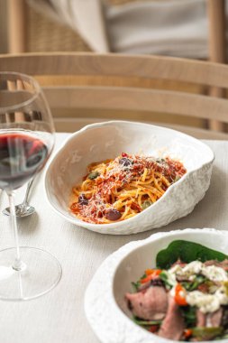Bir bardak kırmızı şarapla servis edilen domates soslu, zeytinli ve parmesanlı İtalyan spagettisi.