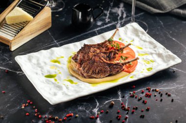 Sulu ızgara kuzu pirzolası, domates dilimleri ve zeytinyağı karışımı ile servis edilir. Zarif beyaz tabakta koyu mermer arka planda.