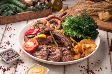 Büyük yuvarlak bir tabakta servis edilen çeşitli ızgara et, sebze, ot ve kızarmış patates.