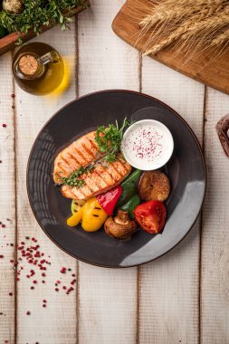 Sulu ızgara somon filetosu. Taze sebze, sos ve siyah bir tabakta otlarla servis ediliyor.