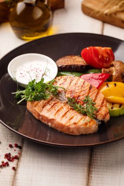 Sulu ızgara somon filetosu. Taze sebze, sos ve siyah bir tabakta otlarla servis ediliyor.