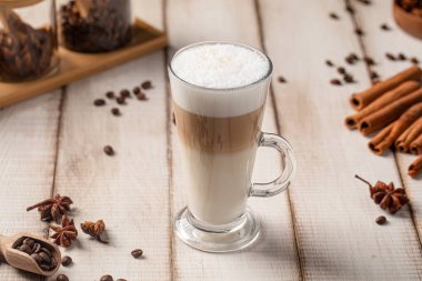 Kahve çekirdekleri ve baharatlarla dolu, ahşap bir masada uzun bir bardakta servis edilen kremalı latte kahve.