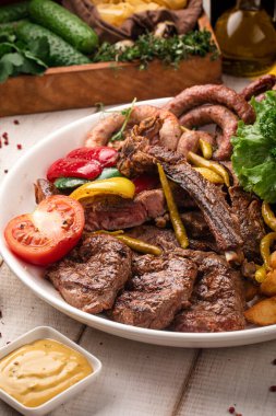 Büyük yuvarlak bir tabakta servis edilen çeşitli ızgara et, sebze, ot ve kızarmış patates.