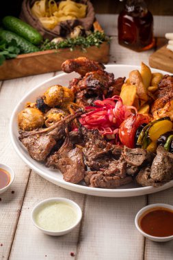 Büyük yuvarlak bir tabakta servis edilen çeşitli ızgara et, sebze, ot ve kızarmış patates.